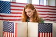 © Tekin - Medium shot, high viewpoint, young, red-haired woman voting at booth during an American election. Sides of booth visible. US flag in background