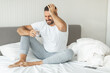 © Prostock-studio - A man sits on a bed, holding a glass of water in one hand and placing the other on his head. His expression is one of pain and discomfort.