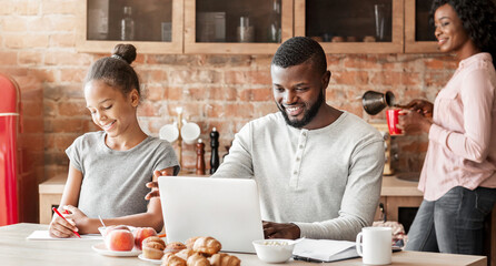 Wall Mural - Happy afro family having breakfast together in kitchen, dad surfing internet on laptop, making plans for weekend, copy space