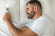 © Prostock-studio - A man lies in bed, propped up on one elbow, his head resting on a pillow. He is looking at a smartphone with his right hand while his left arm is resting on the white bed sheet.