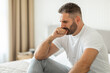© Prostock-studio - A bearded man sits on a bed in a home setting, looking down with a concerned expression, one hand resting on his chin. He wears a white t-shirt and gray pants.
