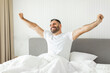 © Prostock-studio - A man in a white t-shirt is sitting up in bed, stretching his arms out to the sides and smiling. He is looking up towards the ceiling as he stretches. The bed has white sheets and a grey headboard