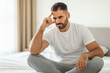© Prostock-studio - A man with a beard, wearing a white t-shirt and grey sweatpants, sits cross-legged on a bed, his hand resting on his forehead. He appears to be deep in thought, with a serious expression on his face.