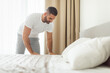 © Prostock-studio - A man in a white t-shirt and gray pants is making his bed in a modern bedroom. He is standing on the side of the bed and has just finished tucking the sheets. White curtains are in the background.