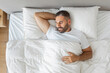 © Prostock-studio - A bearded man is sleeping in a bed with white bedding. He is wearing a white t-shirt and has his arm behind his head. The bed has a white duvet and two white pillows, top view