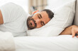 © Prostock-studio - A man with a beard sleeps peacefully on his side, his head resting on a white pillow and his arm outstretched. The bedding is all white and the room appears to be a bedroom.