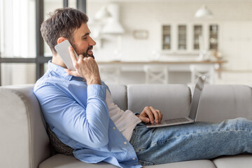 Poster - A man wearing a blue button-down shirt and jeans sits on a gray couch in a living room. He is talking on a white smartphone and working on a laptop computer.