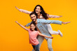 © Prostock-studio - A happy family of three is playing airplane on a yellow background. The father is holding the mother on his back while the daughter is holding on to his shoulders. They are all smiling