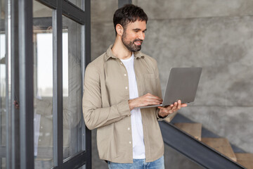 Aufkleber - A man with a beard is standing near a glass door in a modern office, working on a laptop. He is wearing a light brown shirt over a white t-shirt and blue jeans. He is smiling and looks focused