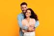 © Prostock-studio - A man with a beard is embracing a woman with long dark hair. They are both smiling and looking directly at the camera. They are standing in front of a solid yellow background.