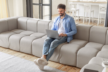 Poster - A man sits on a large, modern, gray sofa, working on his laptop computer. The man is wearing blue jeans and a light blue shirt over a white undershirt, high angle view