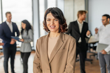 Aufkleber - A woman in tan blazer stands confidently in the middle of a modern office space, with a group of colleagues blurred in the background. She smiles warmly, radiating self-assurance and professionalism.