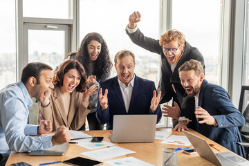 Aufkleber - A group of diverse business colleagues, including both men and women, gather around a laptop in a modern office. They are all smiling and laughing excitedly, with several arms raised in the air.