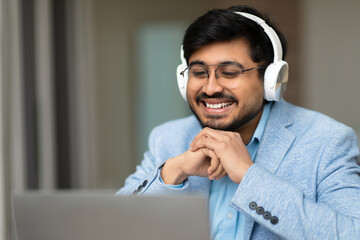 Aufkleber - Indian man is smiling while wearing headphones and looking at a laptop screen. He is seated indoors with natural light coming in through a window to the left of the frame.