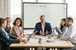 © Prostock-studio - A team of five business professionals sit around a large wooden table in a modern office. The man at the head of the table is presenting a large graph on the screen behind him
