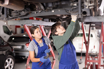 Wall Mural - Young guy and young woman mechanics in uniform repairing underbody of car in car service station