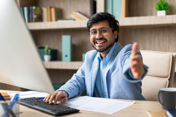 Aufkleber - Indian businessman in a blue suit smiles and gives a thumbs up while sitting at his desk and working on his computer. There are papers on his desk, and a bookshelf is visible behind him.