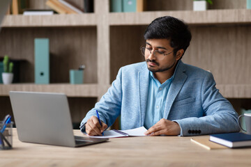 Aufkleber - Indian man in a blue blazer and glasses sits at a wooden desk, writing in a notebook with a blue pen. His laptop is open in front of him, and there are a few other notebooks and a mug on the desk.