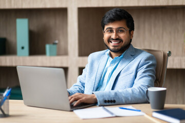 Aufkleber - A young Indian man in a blue suit sits at a desk in an office, smiling as he types on a laptop computer. He has a cup of coffee beside him, and there are papers and a notepad on the desk.