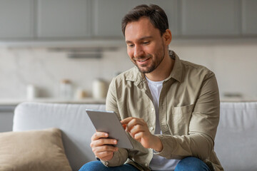 Poster - A man sits on a couch in a living room, smiling as he uses a tablet computer. He is wearing a beige button-down shirt and blue jeans.