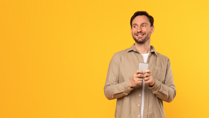 Poster - A man in a tan shirt stands against a yellow background, smiling as he looks at copy space while holding a smartphone in his hands.