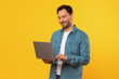 © Prostock-studio - A man wearing a blue denim shirt stands against a yellow background, smiling as he uses a laptop computer.