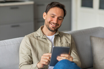 Poster - A man sits on a gray couch in a living room, smiling while using a tablet. He is wearing a beige button-down shirt and blue jeans. He is looking down at the tablet.