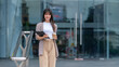 © wattana - A young businesswoman stands confidently in front of a modern glass building, holding a cup of coffee and a clipboard.