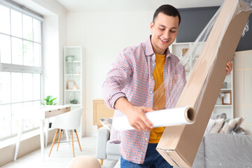 Sticker - Young man wrapping cardboard box with stretch film at home