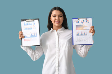 Wall Mural - Young beautiful businesswoman holding clipboard with graphs on blue background