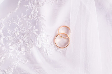 Two gold wedding rings are lying on a white embroidered veil. white satin background. top view. Layout for a wedding opening, invitation.