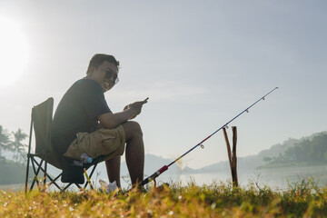 Young Asian man is sitting on a lawn chair by a lake, fishing and looking at his phone.