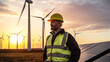 © Mudha Studio - A male engineer in a high-visibility vest and yellow hard hat, standing among wind turbines and solar panels during sunset, highlighting renewable energy