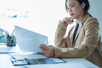 Wall Mural - Businesswoman sitting at desk on couch in workplace or at home working on laptop and analyzing data on charts and graphs and writing on papers to make business plan 