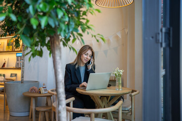 Busy businesswoman owner of modern cafe working at laptop, smiling talking on phone sits at table. Relaxed woman analyzing sales seen on computer screen while make cellphone calls waiting coffee order