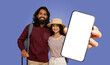 © Prostock-studio - A smiling Indian couple, wearing casual clothing and holding luggage, stand in front of a blank smartphone screen held by woman hand, appears to be ready for travel and excited about their journey.