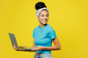 Wall Mural - Side view young Latin IT woman she wears blue top t-shirt headband casual clothes hold use work on laptop pc computer look aside on area isolated on plain yellow background studio. Lifestyle concept.