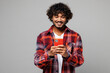 © ViDi Studio - Young happy smiling Indian man he wears red shirt casual clothes hold in hand use mobile cell phone browsing internet online isolated on plain grey color background studio portrait. Lifestyle concept.