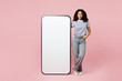 © ViDi Studio - Full body little kid teen girl wearing blue t-shirt casual clothes stand near big huge blank screen mobile cell phone smartphone with area isolated on plain pastel pink background. Childhood concept.