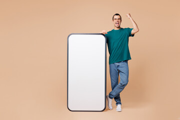 Wall Mural - Full body young middle eastern man wear blue t-shirt casual clothes big huge blank screen mobile cell phone smartphone with workspace area do winner gesture isolated on plain pastel beige background