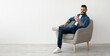 © Prostock-studio - Cheerful young Arab man having online business meeting on smartphone, sitting in cozy armchair near white wall, banner design with free space. Eastern guy communicating remotely on mobile device