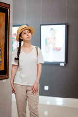 Wall Mural - Historic education and museum day. Pretty young Caucasian woman student wearing straw hat admiring paintings in the gallery. Vertical. Concept of cultural education