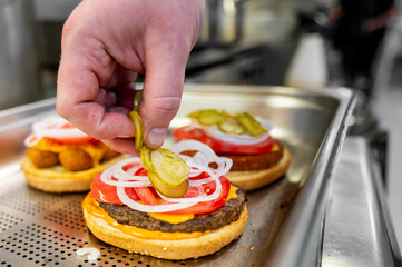 Wall Mural - Close-up of a hand placing pickles on a freshly made burger with cheese, onions, and tomatoes. The background is blurred, emphasizing the burger assembly process. Ideal for food and culinary themes.