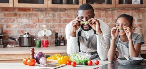 Wall Mural - Black funny father and daughter making mustaches with lettuce, having fun while cooking at kitchen, panorama with empty space