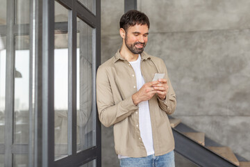 Poster - A man with a beard, wearing a tan shirt over a white t-shirt and jeans, smiles as he scrolls through his smartphone. He is standing next to a modern building with a glass door and concrete walls.