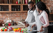 © Prostock-studio - Lovely african couple using digital tablet while cooking vegetarian meal, copy space