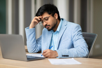 Aufkleber - Indian young man with a beard is sitting at a desk, wearing a light blue blazer and glasses, focusing intently as he works. He has his hand on his forehead and is writing on a paper with a pen