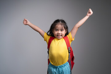 Wall Mural - Asian young girl in a yellow shirt and red backpack is jumping up and down with her arms raised, studio on grey background.