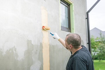 Wall Mural - A man in a black shirt and white gloves uses a paint roller to paint a vertical stripe on a house wall. A window is to his right.