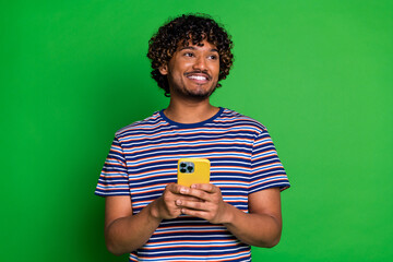 Sticker - Portrait of young curly haired guy in striped t shirt using smartphone looking empty space shopping isolated on green color background